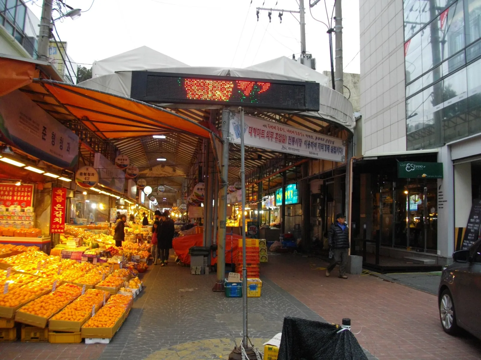夕暮れの済州の東門市場で、果物の屋台と店の看板が並ぶ市場の通り