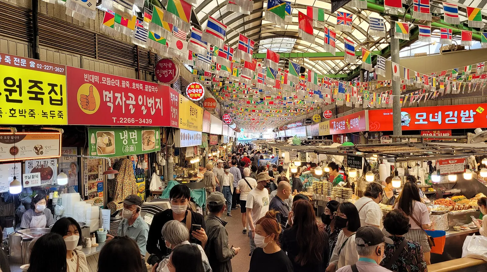ソウル広蔵市場で、吊り下げられた旗と明るい看板の下に並ぶ、にぎやかな食べ物通り
