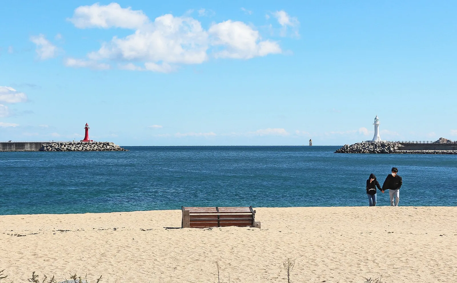 Sandy Sokcho beach with a bench, two distant visitors and a red lighthouse on the breakwater